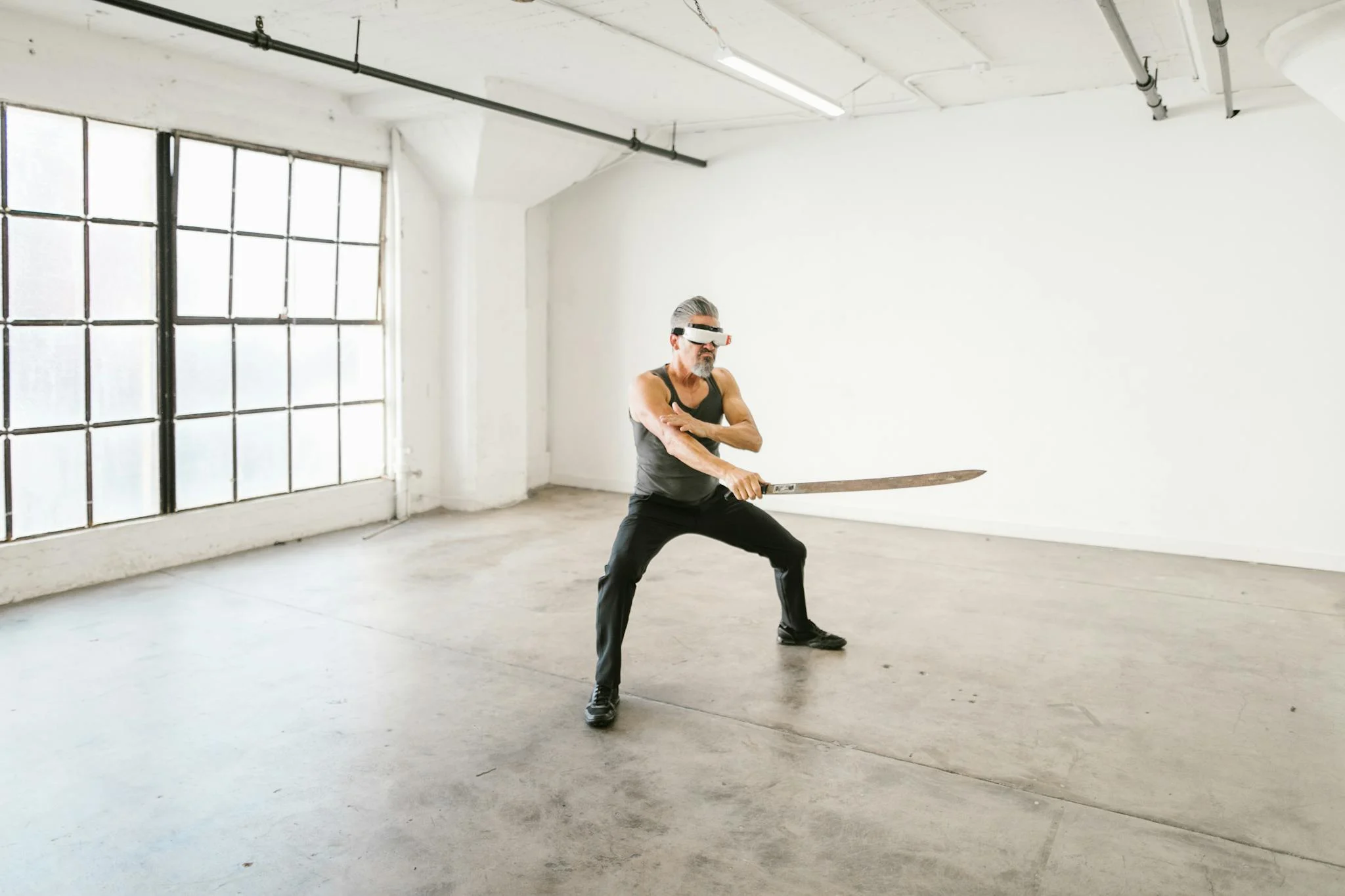 An adult man practicing sword techniques while wearing a VR headset indoors, minimalist setting.