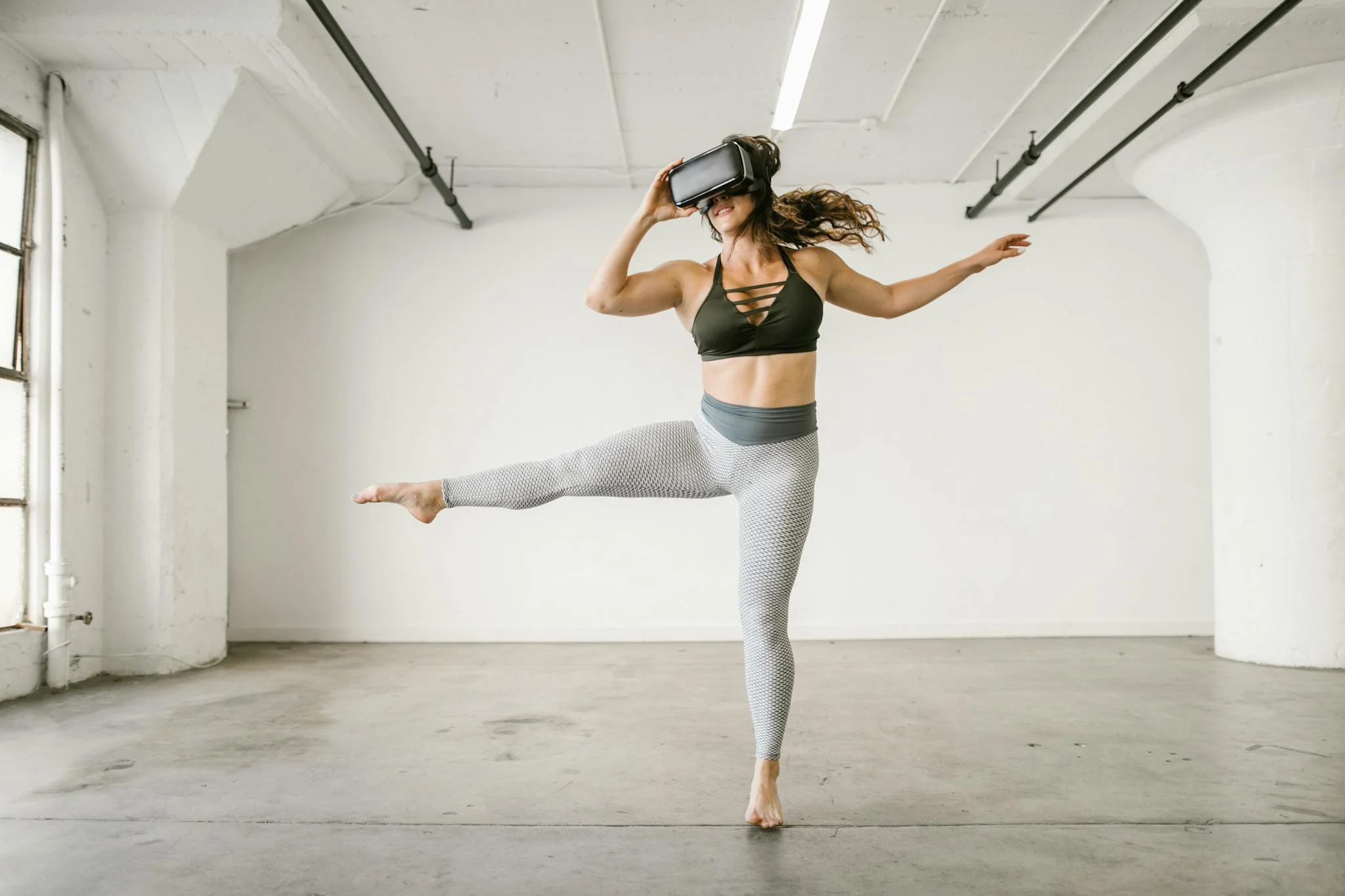 Woman in athletic attire explores virtual reality using a VR headset while dancing in a modern studio.
